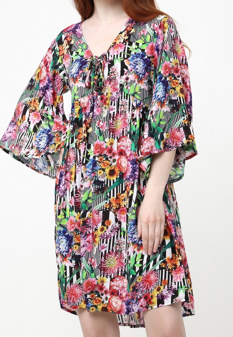Femme portant une robe fleurie multicolore avec des rayures diagonales noires et blanches, des manches 3/4 larges et un détail noué au niveau du décolleté.