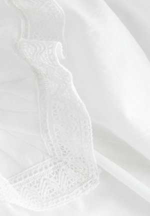 Tissu blanc avec une bordure en dentelle blanche complexe et un bord ondulé, mettant en valeur des détails fins et une texture douce.