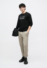 Mustade varrukatega särk logoga "EA7 Emporio Armani", beežid kitsenevad püksid ja mustad tossud, käes must kott. Minimalistlik disain.