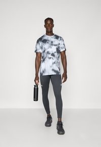 Sportliches Outfit mit einem grauen Batik-T-Shirt, gerippten, dunkelgrauen Leggings und schwarzen Sneakers, dazu eine schwarze Wasserflasche in der Hand.
