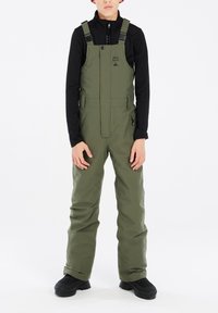 Salopette impermeabile verde oliva con bretelle regolabili, tasca con zip frontale, tasche laterali e design a gamba larga, indossata sopra una maglietta nera a maniche lunghe.