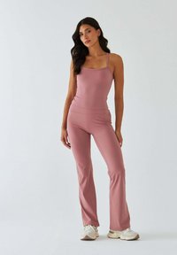 Różowe bezrękawnikowe body w połączeniu z różowymi spodniami rozkloszowanymi, oba wykonane z gładkiej, rozciągliwej tkaniny. Modelka ma na sobie białe trampki.