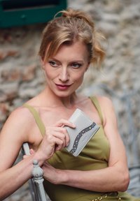 Šedá peněženka s křivkovým designem a barevnými puntíky. Vyrobena z hladkého materiálu, držena v ruce v zelených šatech.