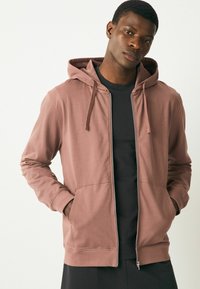 Hooded sweatshirt in dusty rose, made of soft fabric. Features a front zipper, two side pockets, and a relaxed fit, paired with a black top underneath.