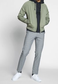 Giacca bomber verde oliva con cappuccio, indossata sopra una felpa nera, abbinata a pantaloni a quadri grigi chiari e sneakers bianche.