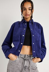 Giacca di velluto a coste blu navy con due tasche sul petto, chiusura a bottoni a pressione e vestibilità rilassata, abbinata a pantaloni grigi con cordoncino.
