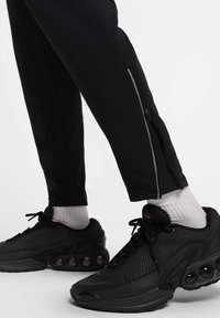 Des joggers athlétiques noirs avec des fermetures éclair aux chevilles sont associés à des baskets noires texturées avec des accents arrondis et des chaussettes blanches. 