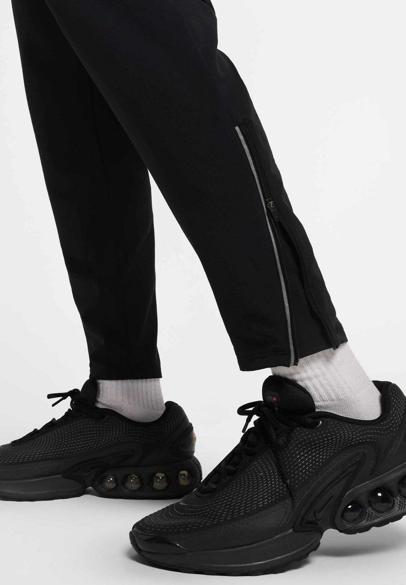 Des joggers athlétiques noirs avec des fermetures éclair aux chevilles sont associés à des baskets noires texturées avec des accents arrondis et des chaussettes blanches. 