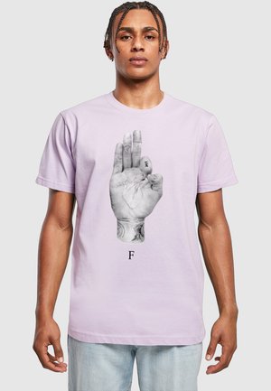 Młody mężczyzna z zaplecionymi włosami ma na sobie jasnofioletowy t-shirt z czarno-białą grafiką ręki pokazującą literę F w amerykańskim języku migowym.