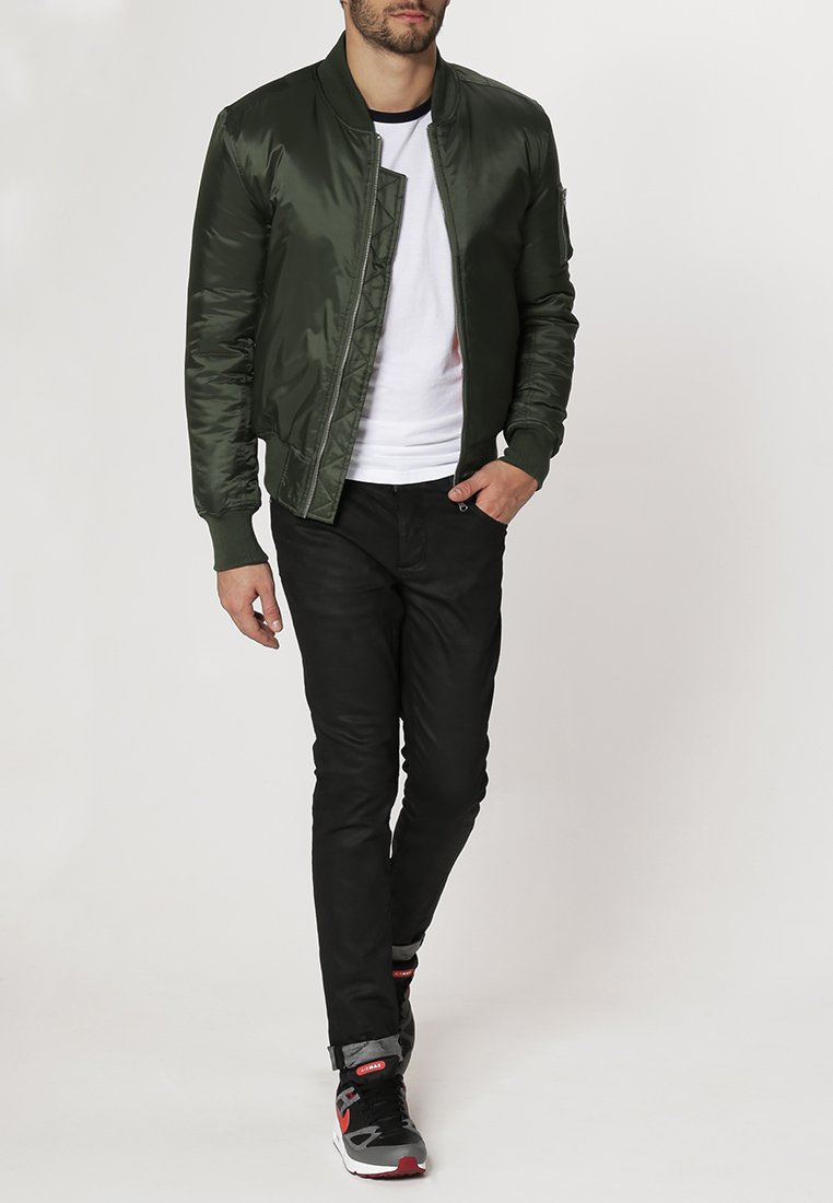Mann geht, trägt eine dunkelgrüne Bomberjacke, ein weißes T-Shirt, schwarze Hose und schwarze, graue und rote Sneakers.
