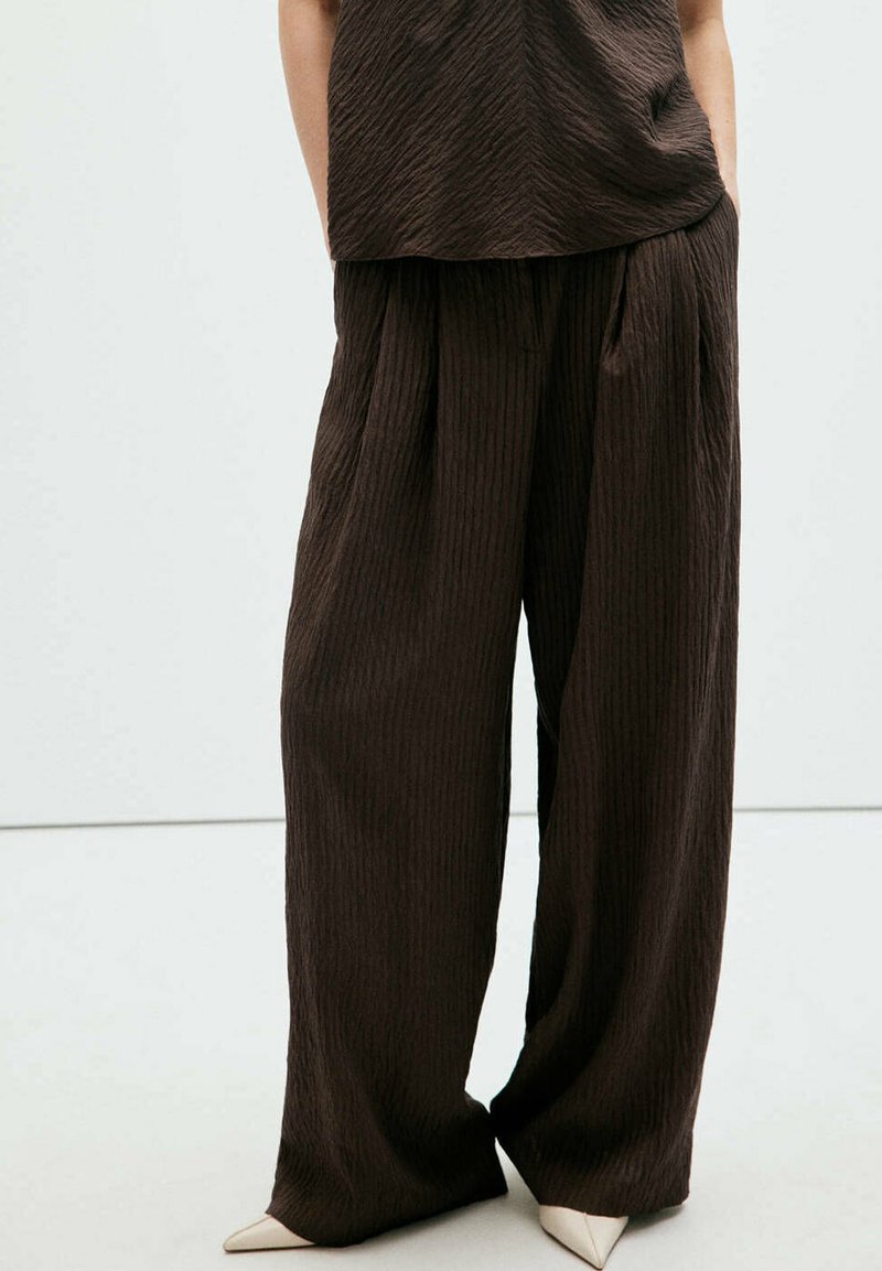 Personne portant un pantalon large marron foncé texturé et un haut assorti, associé à des chaussures à talons aiguilles beiges pointues.