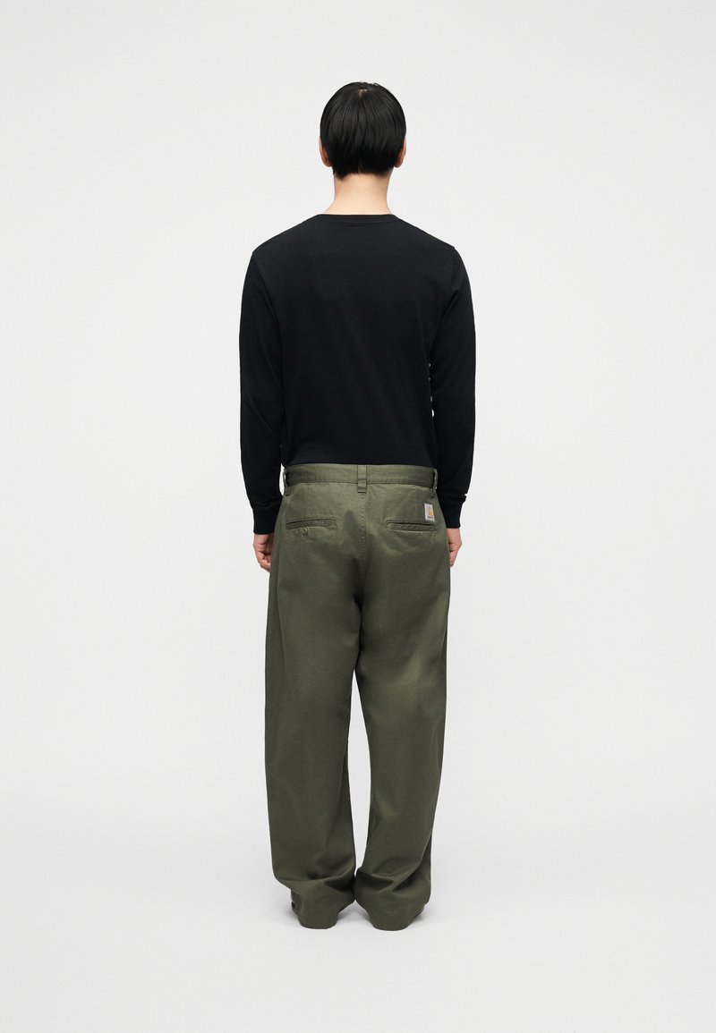 Chemise noire à manches longues associée à un pantalon ample de couleur vert olive, présentant un design simple, une texture douce et des éléments métalliques minimaux.