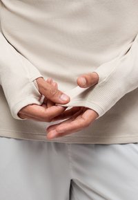 Χέρια που ρυθμίζουν τις τρύπες για τους αντίχειρες στα μανίκια ενός υφασμάτινου, ανοιχτού μπεζ μακρυμάνικου πουκαμίσου πάνω από ανοιχτό γκρι παντελόνια.