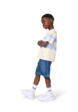 Garçon bras croisés portant un t-shirt rayé, un short en jean, des baskets blanches et des chaussettes blanches avec le logo "BOSS", se tenant sur un fond blanc.