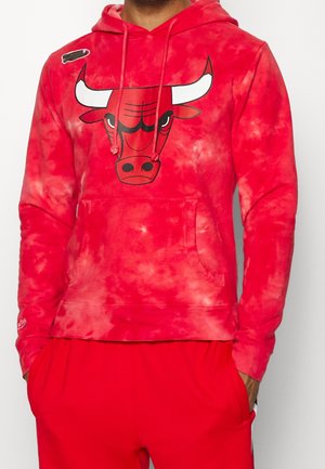 Person iført en rød tie-dye hættetrøje med et stort tyr-hoved grafik og matchende røde bukser, hænder i lommerne, mod en ensfarvet baggrund.
