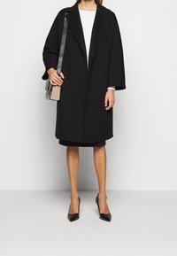Schwarzer Wollmantel mit breitem Schal-Kragen und tiefen Schultern. Kombiniert mit einem figurbetonten schwarzen Kleid und spitzen schwarzen High Heels. Karierter Beutel in der Hand.