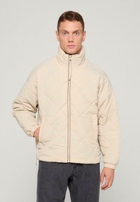 Beżowa pikowana kurtka z wysokim kołnierzem, zamek błyskawiczny z przodu i elastycznymi mankietami. Miękka tkanina i detal w romby. Casualowy design.