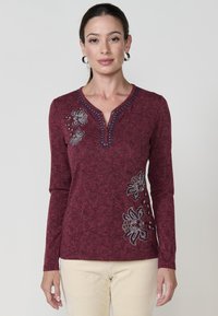 Sieviete, kura valkā burgundijas krāsas garām piedurknēm krekliņu ar ziedu izšuvumu un pogātu V-veida izgriezumu, apvienota ar gaiši bēšām biksēm uz vienkārša fona.