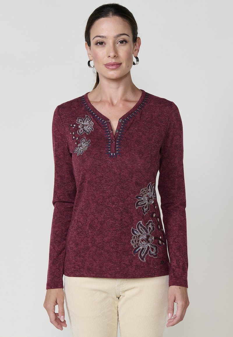 Sieviete, kura valkā burgundijas krāsas garām piedurknēm krekliņu ar ziedu izšuvumu un pogātu V-veida izgriezumu, apvienota ar gaiši bēšām biksēm uz vienkārša fona.