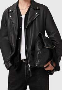 Giacca da moto in pelle nera con hardware argentato, frontale con zip, colletto rialzato e due tasche laterali. Abbbinata a una camicia bianca e a una borsa nera.