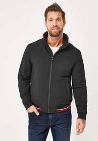 Schwarze Bomberjacke mit Reißverschluss, ausgestattet mit einem Stehkragen und roten Akzenten sowie gerippten Bündchen. Modell kombiniert mit blauen Jeans.