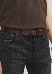 Brun læderbælte med en rektangulær sort metalspænde, båret over mørke denimbukser med en tekstureret finish og en tætsiddende pasform.