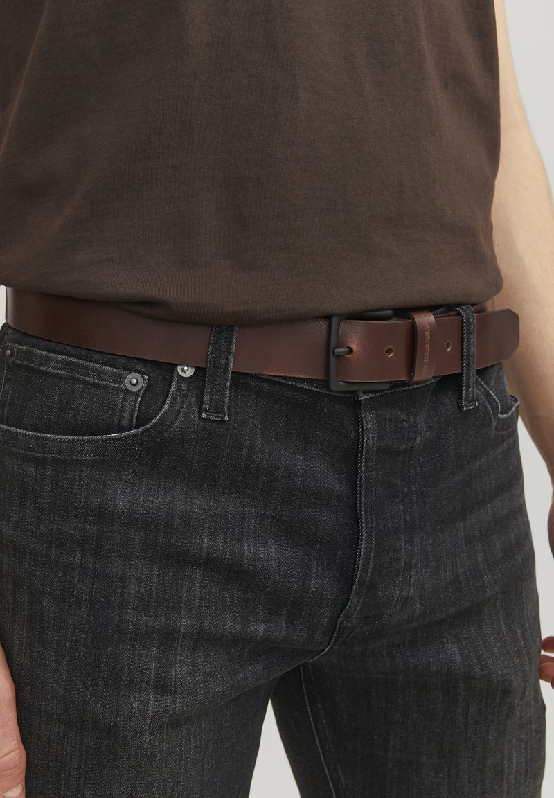 Brun læderbælte med en rektangulær sort metalspænde, båret over mørke denimbukser med en tekstureret finish og en tætsiddende pasform.