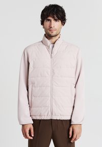 Mann mit dunklem Haar, der eine hellrosa gesteppte Zip-Jacke und braune Hose trägt, steht vor einem schlichten weißen Hintergrund.