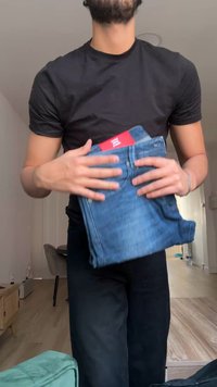 Man die een zwart shirt en broek draagt, houdt opgevouwen blauwe spijkerbroek met een rood label vast in een moderne woonkamer.