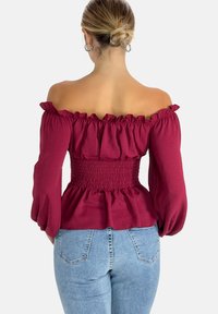 Burgunderfarbenes Off-Shoulder-Oberteil mit Rüschensaum am Ausschnitt und an den Ärmeln, gerafftem Taillenbereich und ausgestellt geschnittenem Saum. Kombiniert mit hellblauen Jeans mit Gesäßtaschen.