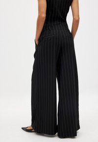 Široke črne hlače z navpičnimi belimi črtami. Narejene so iz gladkega materiala, vsebujejo zadnje žepe in imajo prilegajoč dizajn pasu.