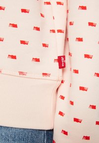 Světle růžová mikina se repetitivním červeným vzorem stylizovaných vlajek. Obsahuje červenou nášivku Levi's na boku a detaily s žebrováním na manžetách.