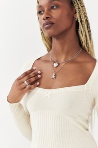 Haut crème côtelé avec encolure carrée, agrémenté d'un accent en forme de nœud blanc. Colliers superposés en argent avec des pendentifs en forme de cœur et d'ours en peluche. Bague en argent.