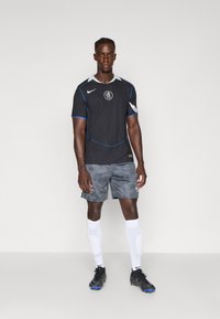 Schwarzes Sporttrikot mit blauen Akzenten, Logo in der Mitte, kombiniert mit gemusterten schwarzen Shorts, weißen Fußballsocken und schwarzen Fußballschuhen.