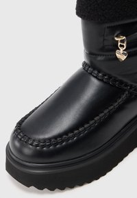Bota de invierno de cuero negro con suela gruesa, costuras visibles, puño de pelaje y pequeño charm en forma de corazón dorado adjunto cerca de la parte superior.