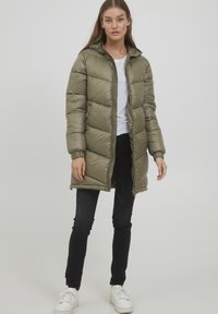 Manteau matelassé vert olive avec capuche, doté d'une fermeture éclair, de poignets élastiqués et d'une texture rembourrée. Porté par-dessus une chemise blanche et un pantalon noir.