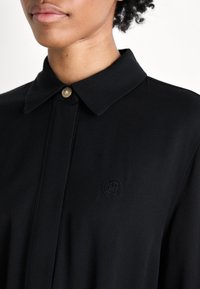 Primo piano di una persona che indossa una camicia nera con colletto, con un unico bottone in alto e un logo ricamato sottile sul petto.