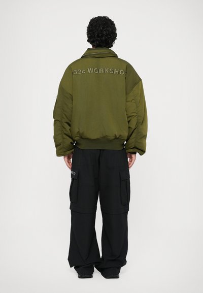 Doudoune vert olive avec le texte "32C WORKSHOP", ourlet côtelé et coutures d'épaule. Associée à un pantalon cargo noir avec poches latérales.