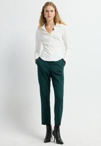 Witte, aansluitende blouse met een kraag, gecombineerd met donkergroene, op maat gemaakte broek. Model draagt zwarte, hoge enkellaarsjes. Gladde stof en gestructureerd ontwerp.