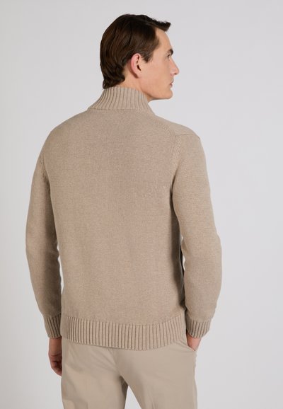 Uomo che indossa un maglione a collo alto a coste beige e pantaloni beige, in piedi con le mani nelle tasche, rivolto di lato e leggermente indietro.