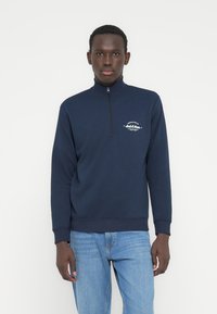 Μπλε ναυτικό φούτερ με φερμουάρ και ψηλό γιακά, ριμπ μανσέτες και τελείωμα. Διαθέτει μικρό λευκό λογότυπο στο στήθος. Μαλακό υλικό με μείγμα βαμβακιού.