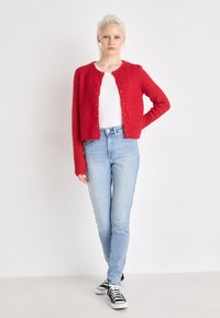 Cardigã vermelho de malha com gola redonda, fecho de botão e mangas longas, combinado com jeans azul claro e ténis pretos.