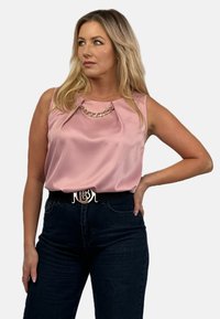 Satynowy różowy bezrękawnik z łańcuszkowym akcentem przy dekolcie, zestawiony z ciemnoniebieskimi dżinsami i czarnym paskiem z metalową klamrą.