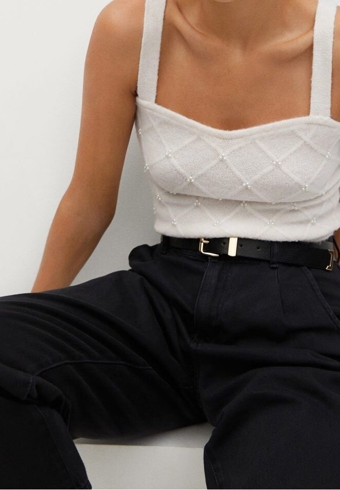 Personne portant un haut sans manches en tricot blanc avec des détails en perles et un pantalon noir taille haute avec une ceinture noire, assise.