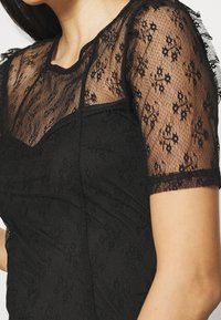 Haut en dentelle noire avec un motif floral transparent, manches courtes, corsage texturé et encolure arrondie.