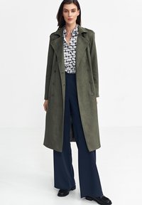 Zielonkawozielony płaszcz typu trench o teksturowanym wykończeniu, podwójnie zapinany, z dużymi klapami, w połączeniu z granatowymi spodniami o szerokich nogawkach i wzorzystą koszulą.