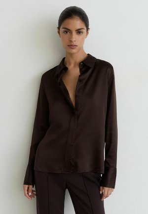 Vrouw met donker haar naar achteren gebonden, gekleed in een losse zwarte blouse met knopen en bijpassende broek, staand tegen een witte muur.