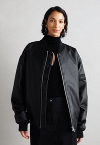 Giacca bomber in finta pelle nera con texture trapuntata, colletto a costine e zip bianca a contrasto. Presenta una vestibilità over e tasche laterali.