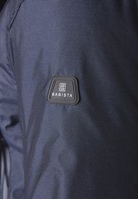 Marineblaue Stoff mit einer strukturierten Oberfläche, die auf der linken Seite einen schwarzen Gummilabel-Patch mit der Aufschrift "BABISTA" aufweist.
