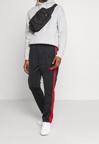 Pantalon de sport noir avec des bandes rouges sur les côtés, associé à un sweat à capuche gris et un sac bandoulière noir. Des baskets blanches complètent la tenue.